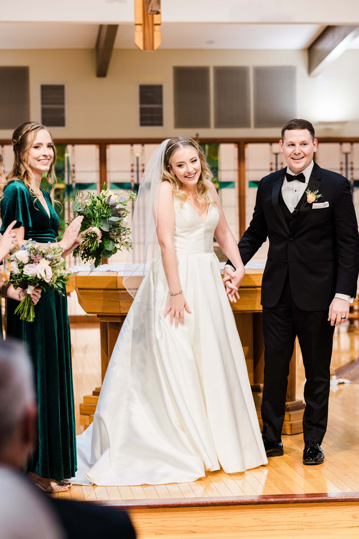 Bride and groom holding hands and smiling, walking down the aisle at Park Farm Winery while a bridesmaid claps on the side in a church.