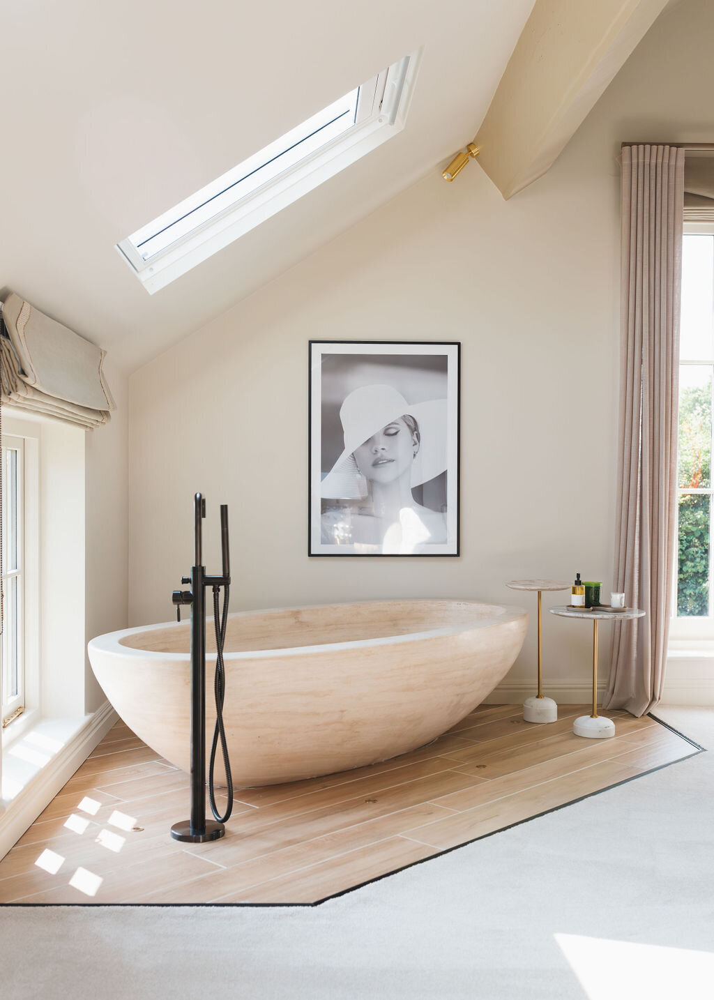 A minimalist neutral bedroom features a large, freestanding wooden bathtub under a skylight. There's a floor-mounted faucet beside the tub, a small side table with decor, and a framed black-and-white photo on the wall. The space is bright with natural light and neutral tones.