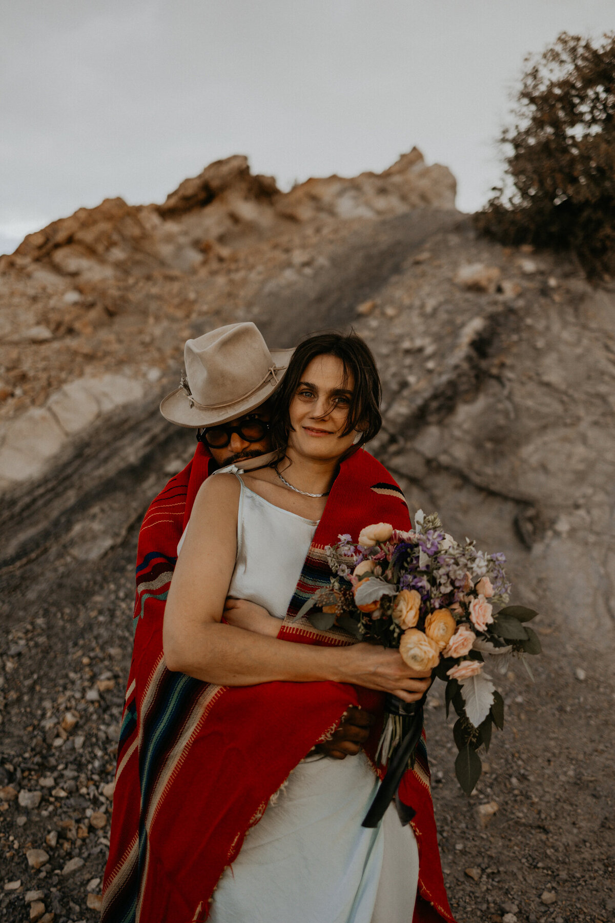 a groom wrapped in a native American blanket hugging his new bride in the desert