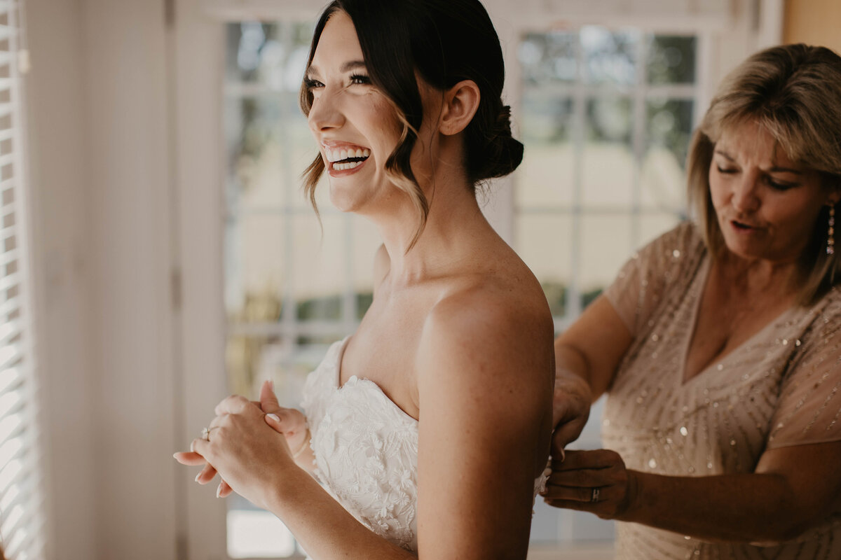 Bride laughing and enjoying her mom zipping her wedding dress. 