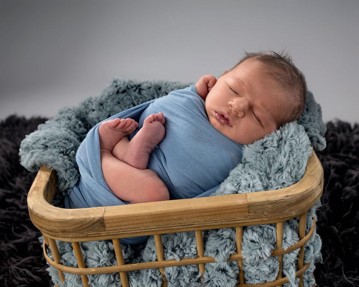 A newborn sleeps in a blue swaddle in a wooden basket