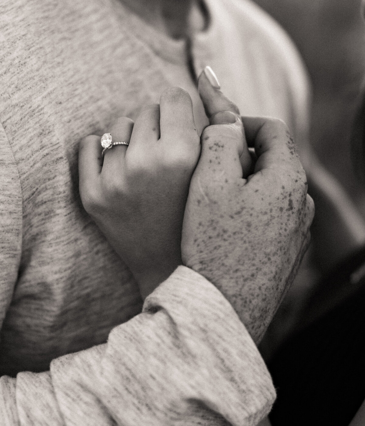 A guy is holding his fiancé's hand close to his chest while she wears a sparkly engagement ring.