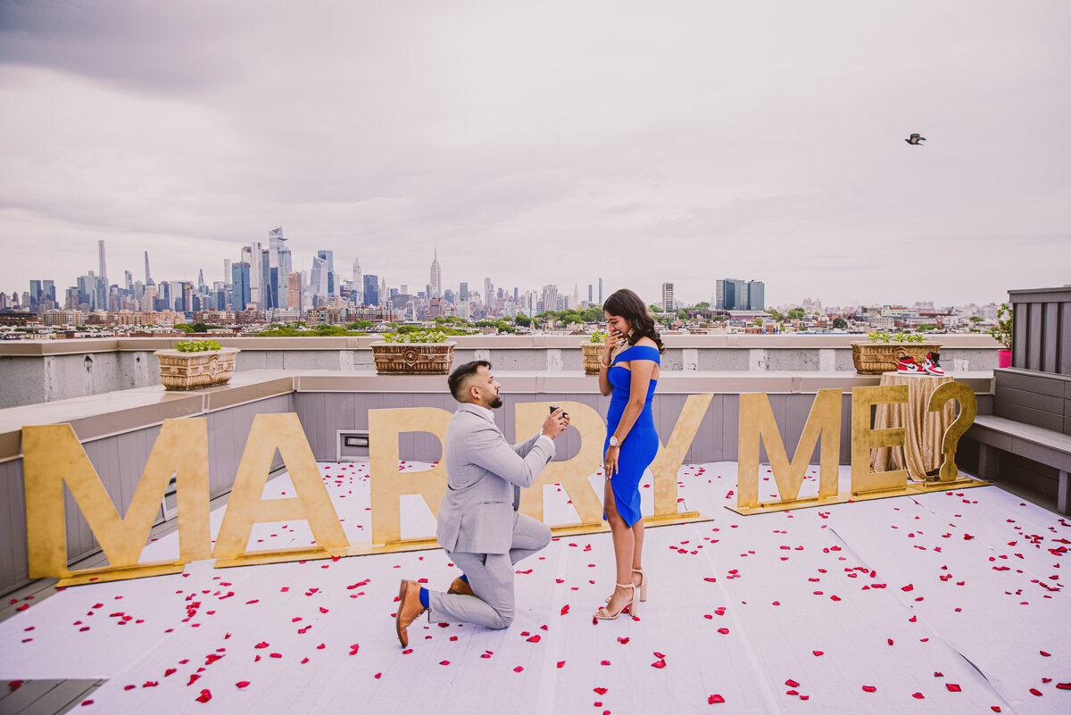 A man proposes to girl while on his knees with the marry me sign in the back on a rooftop in hoboken nj