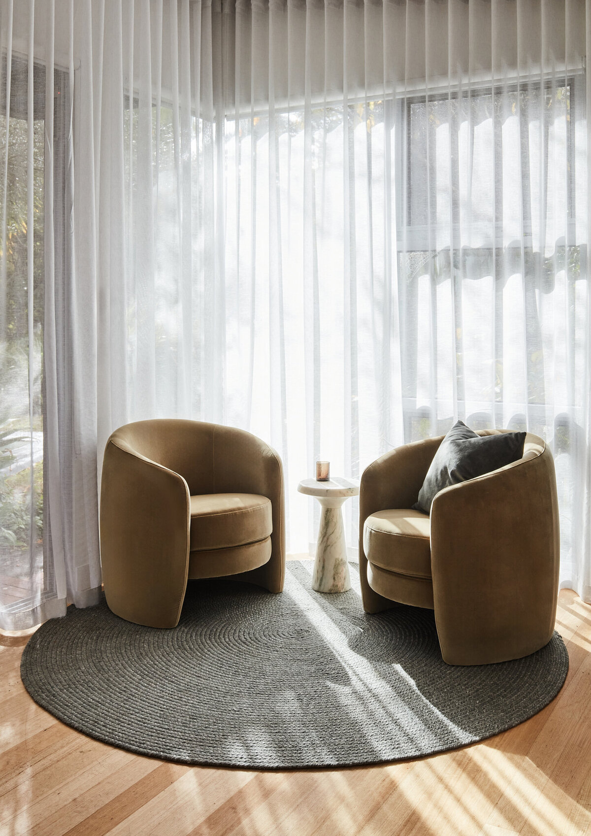 Two tan, upholstered accent chairs with a small marble side table between them on a large, circular gray jute rug. The seating area is in front of a window with sheer white curtains, creating a soft, inviting mood.