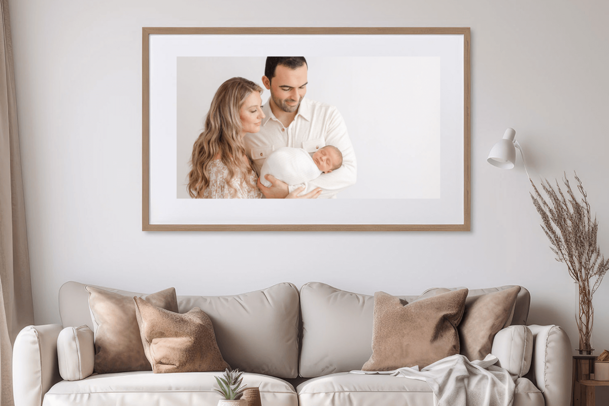 A framed newborn photograph with parents looking down at their sleeping baby. The framed photos is hanging above a neutral couch in a minimalist living room.
