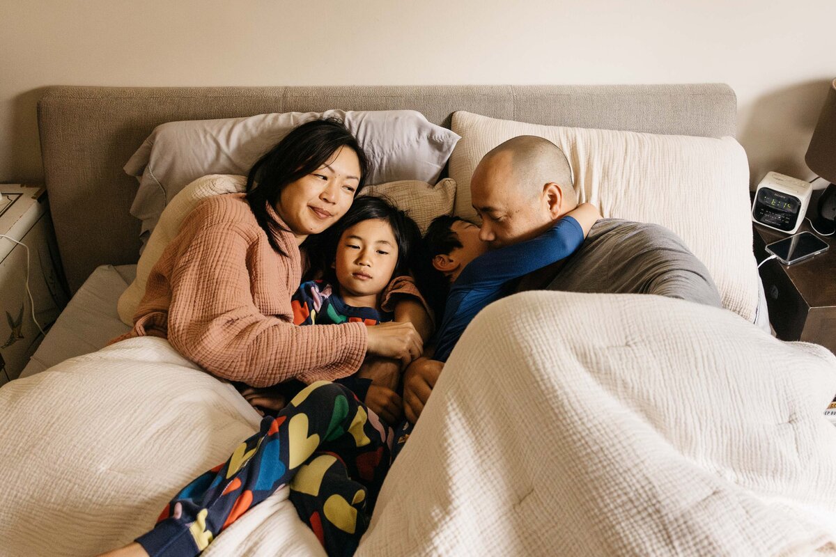 Mother, Husband, and two young boys are snuggling in bed with pajamas on.