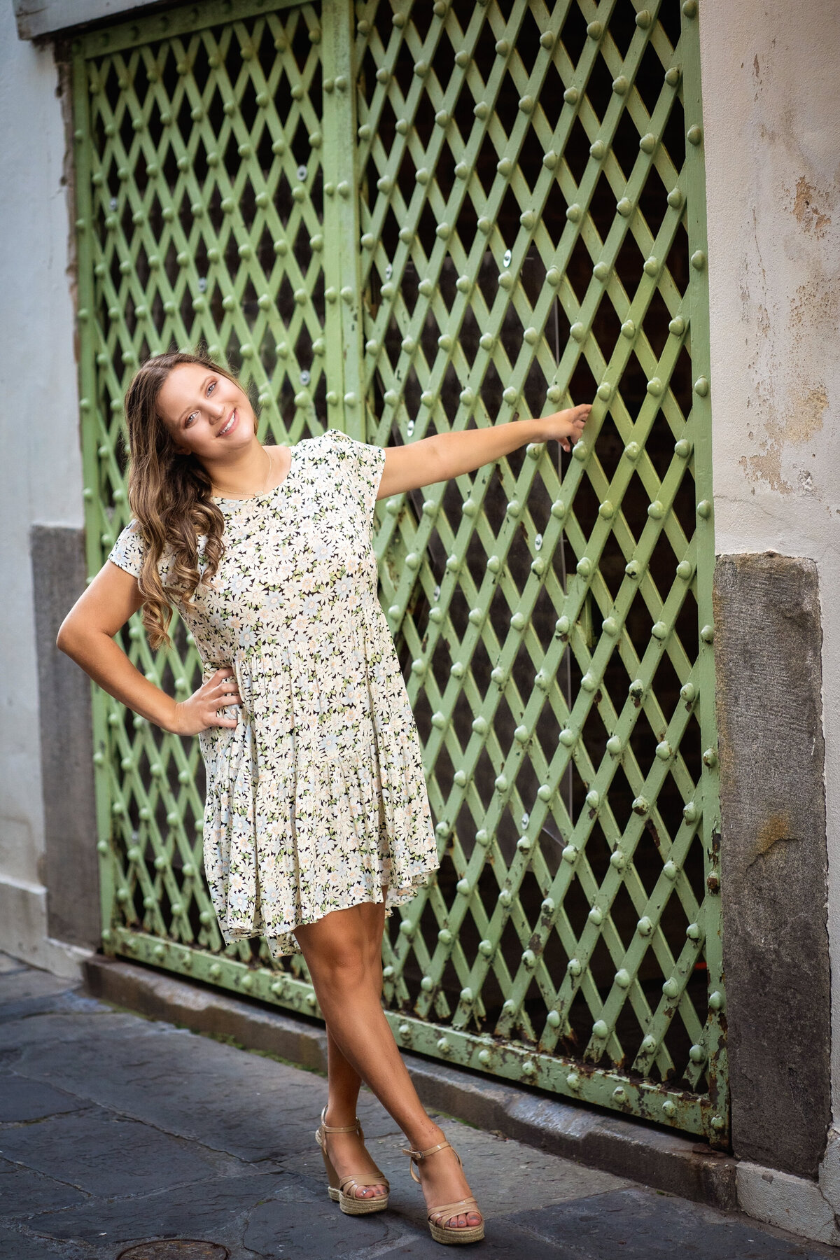 Senior high school girl in a floral patterned dress holding onto a green metal door.  She is wearing nude high heels and smiling at the camera with one hand on her waist.  She has long light brown wavy hair.