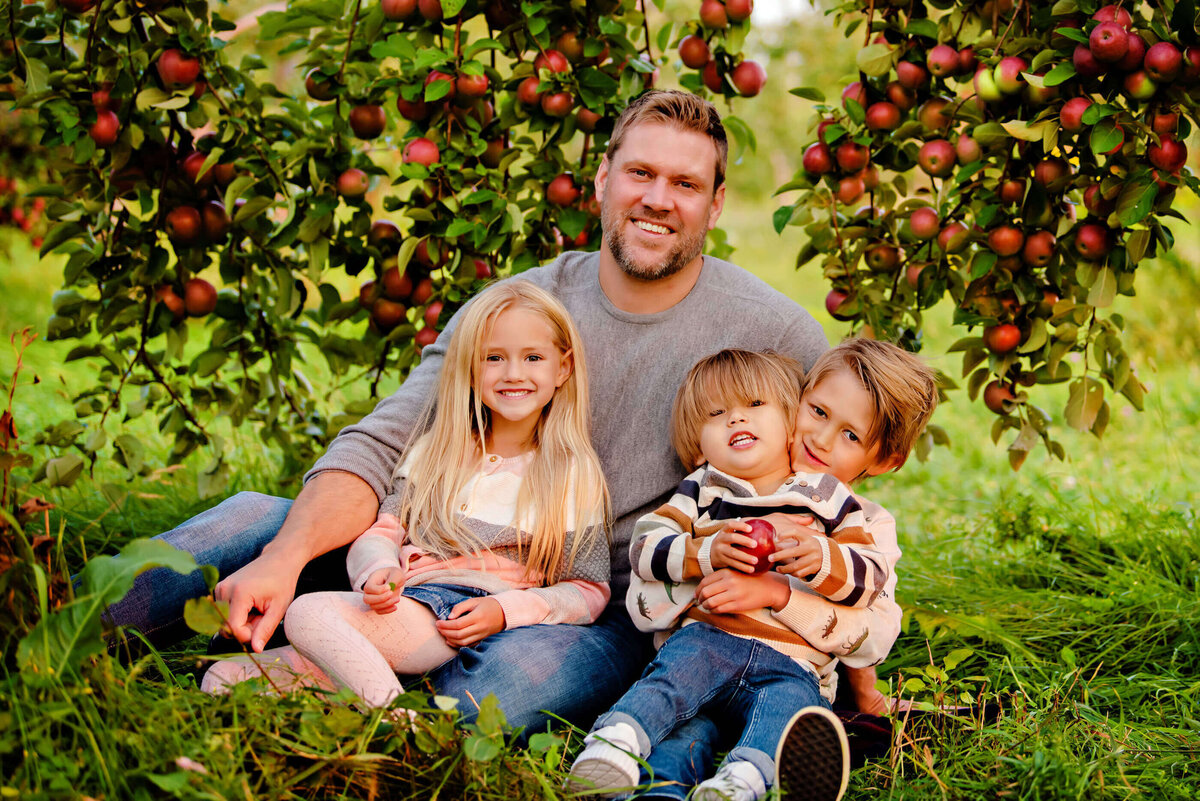 dad sitting on the ground under and apple tree with his two boys and daughter for their fall family photography session in Burlington, ON