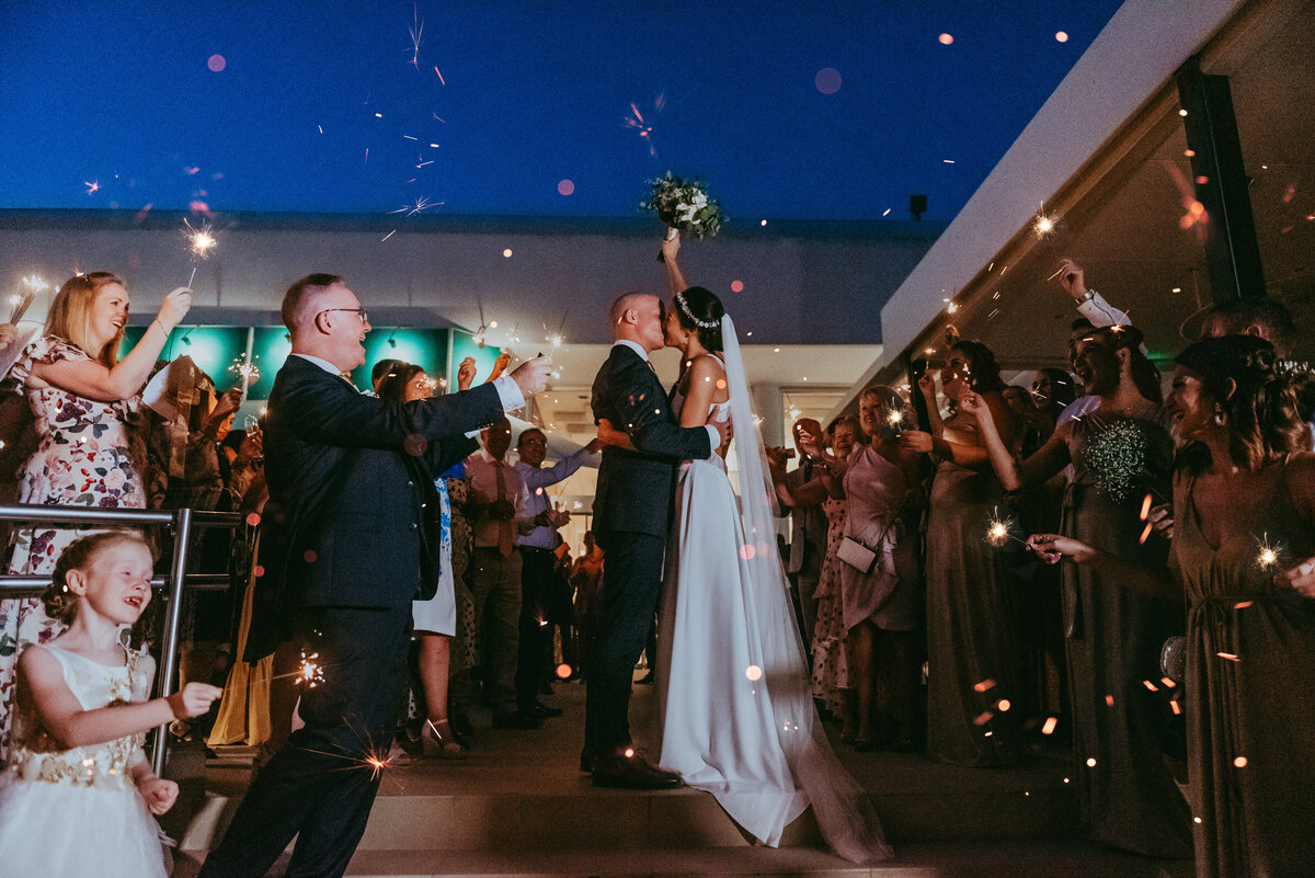 Bride holds bouquet in the air while guests wave sparklers to welcome them to their reception