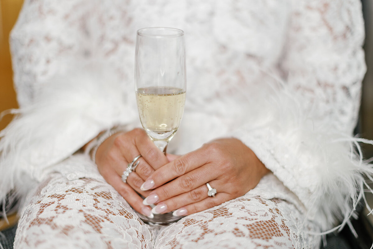 The bride holds a glass of champagne while wearing a lace bridal robe with feather details, embodying luxury and elegance, captured by Claudia Amalia Photography.