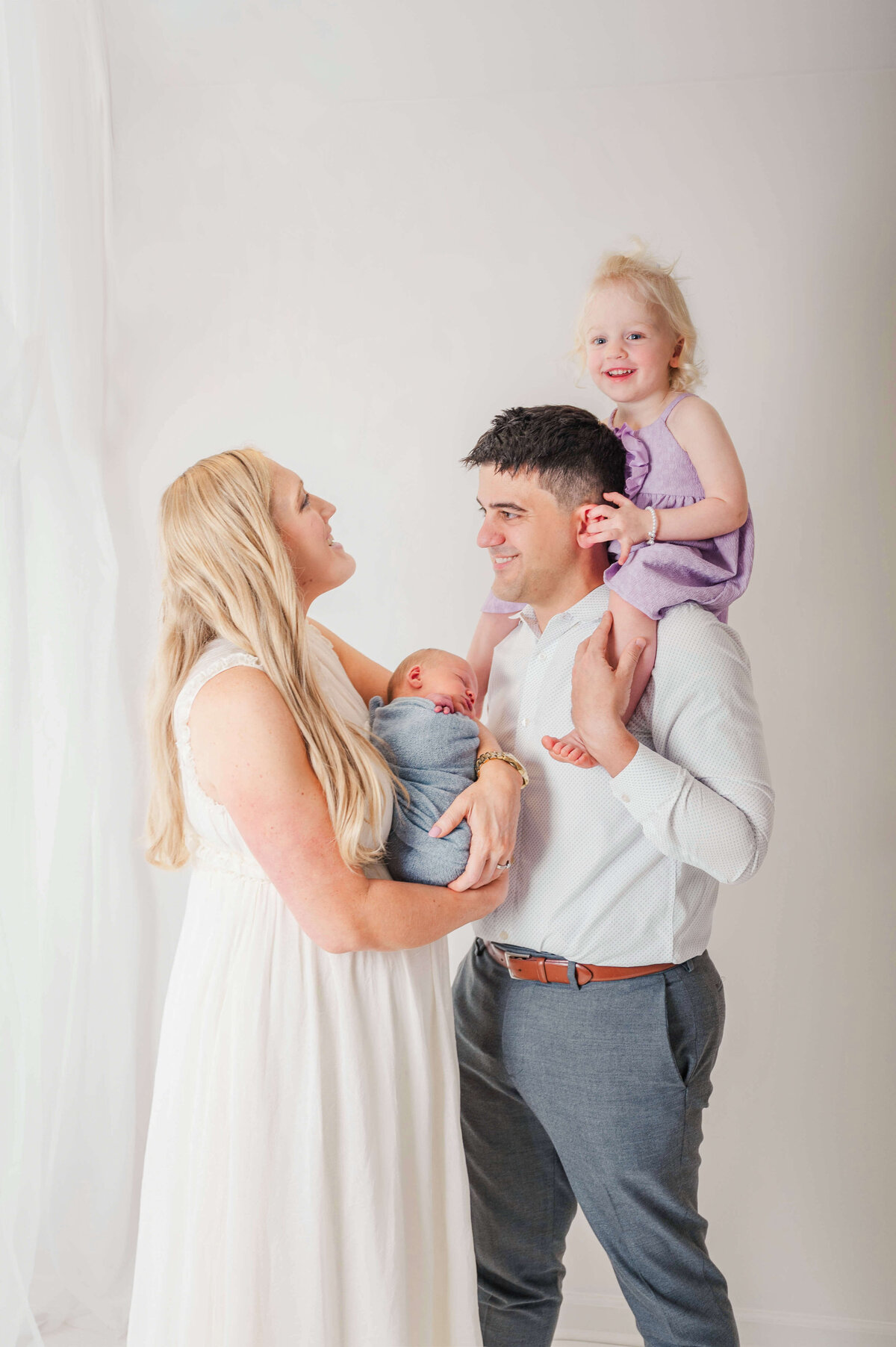 A blond mom holding her newborn and looking up and laughing at her daughter who is sitting on her dad's shoulders.