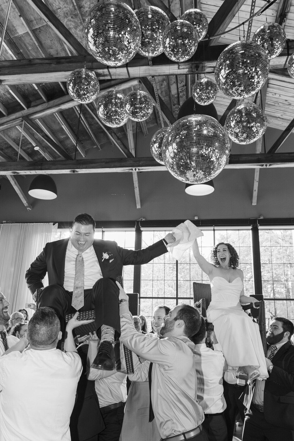 Black and white reception photo of a groom lifted on a chair during the hora beneath disco balls