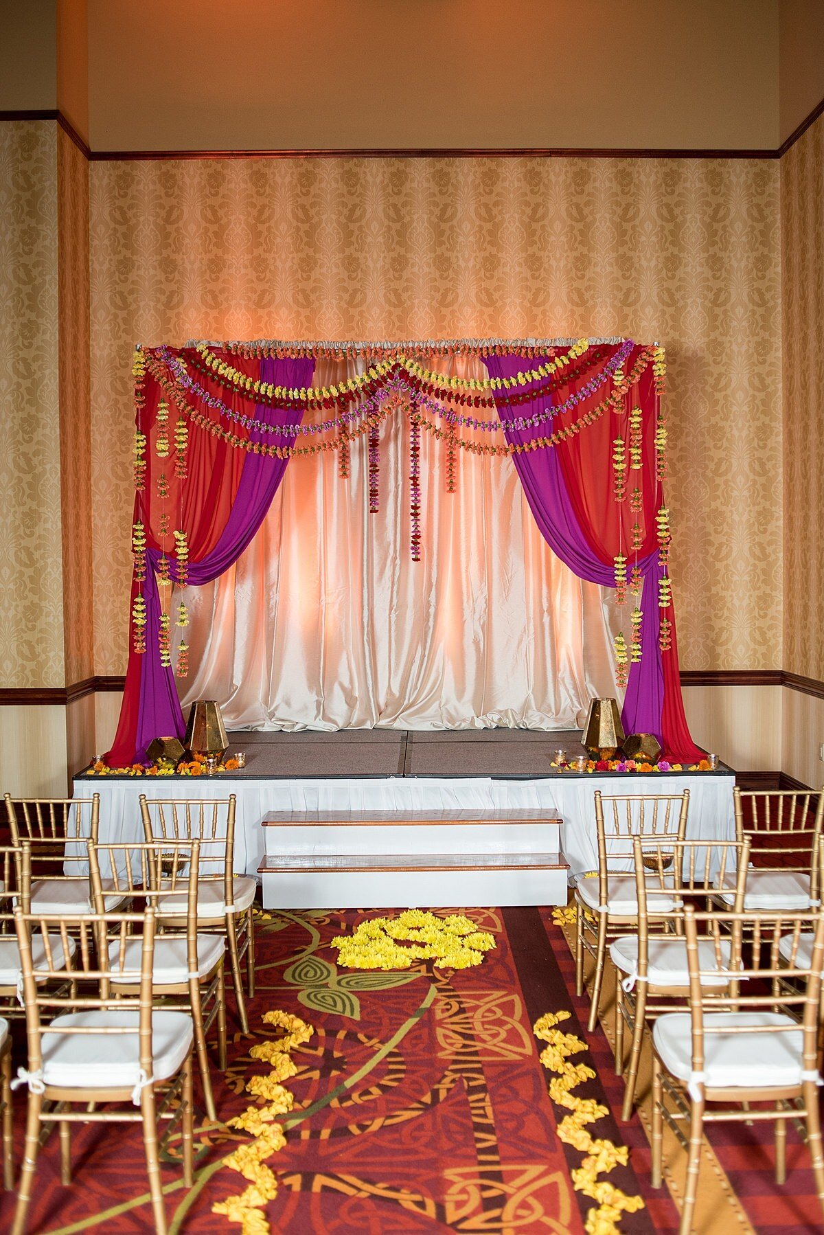 Red and purple mandap decorated with orange, yellow, purple and red floral garlands and yellow flower petal designs down the aisle at a Nashville Indian wedding. Gold chivari chairs with ivory cushions and a red and gold carpet at Embassy Suites Hindu wedding