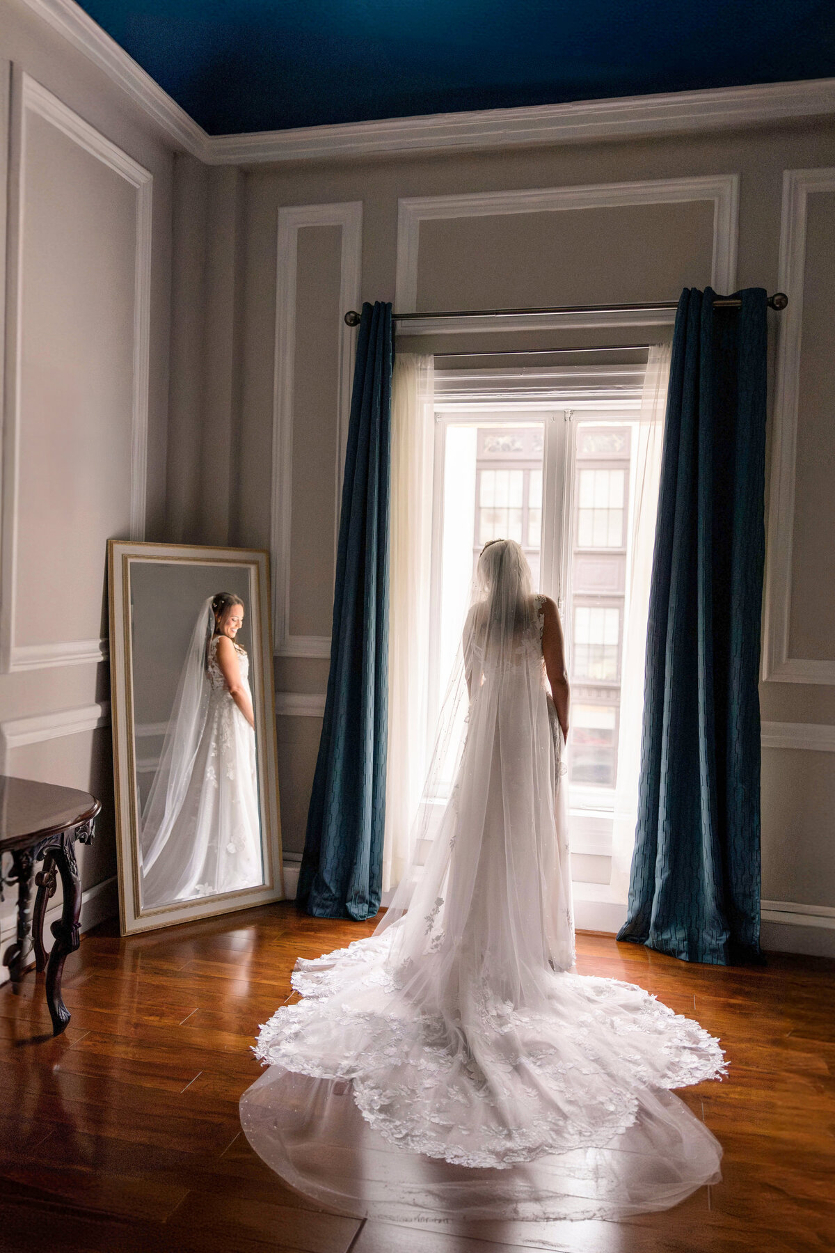 A bride in a long white gown stands facing a large window with blue curtains, her reflection visible in a mirror beside her. The room has dark wooden floors and a high ceiling, creating an elegant setting.