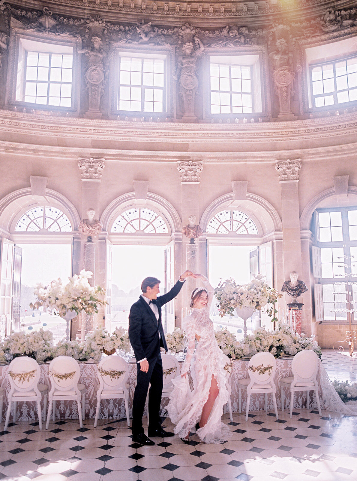 The photo was taken in Paris, France - Chateau Vaux le Vicomte, a top destination wedding venue in France, captured beautifully by photographer Audrey. In addition to capturing the magical moment, you can also add East West Films as videographers for this event. The stunning setting features a bride and groom dancing in an ornate, sunlit room with tall arched windows. The elegant space is adorned with delicate white flowers and statues, creating a romantic ambiance. The groom looks dashing in a black suit, while the bride is radiant in a flowing, lace white dress with a long train. Truly a picture-perfect moment captured in a dreamy location!
