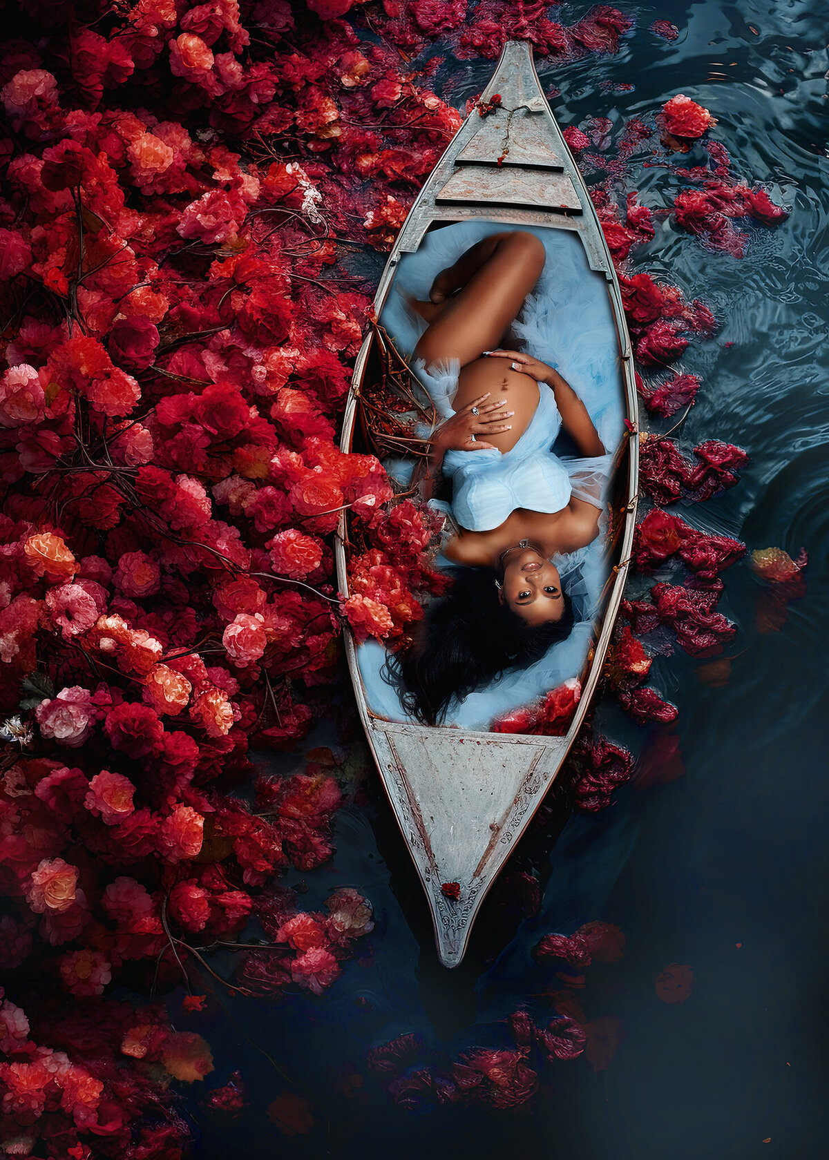 Pregnant woman laying down in a conoe on water with red flowers surrounding the canoe, she is wearing a baby blue gown