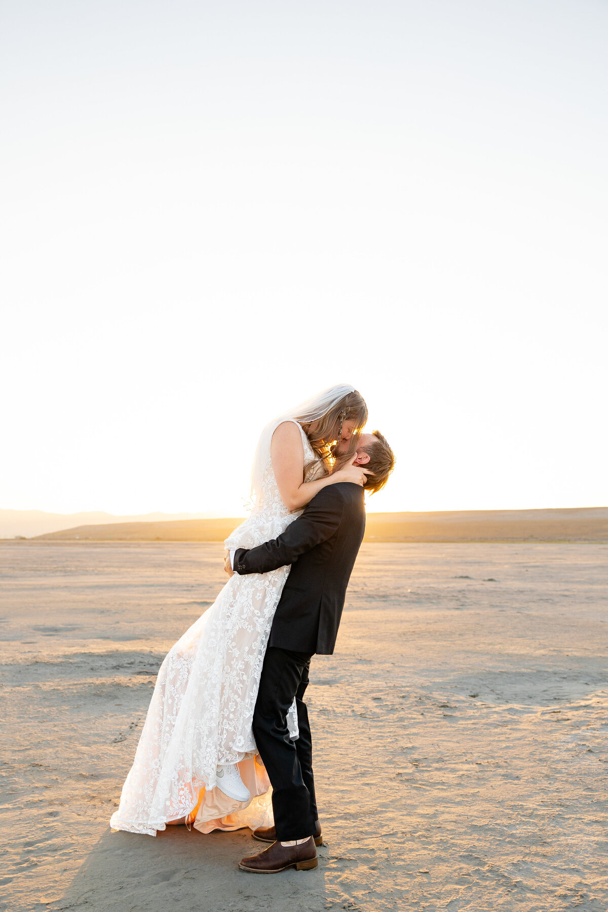 Groom lifting the bride in a joyful embrace, both smiling as the sun rises over the desert landscape.