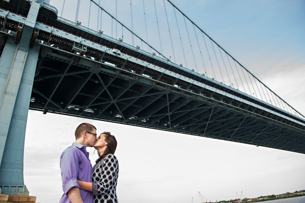 A couple kiss under the ben franklin bridge in philadelphia.