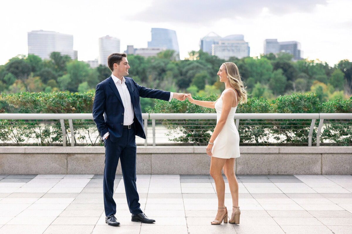 A man in a dark suit and a woman in a white dress stand on a terrace, facing each other and holding hands. The city skyline and greenery are visible in the background.
