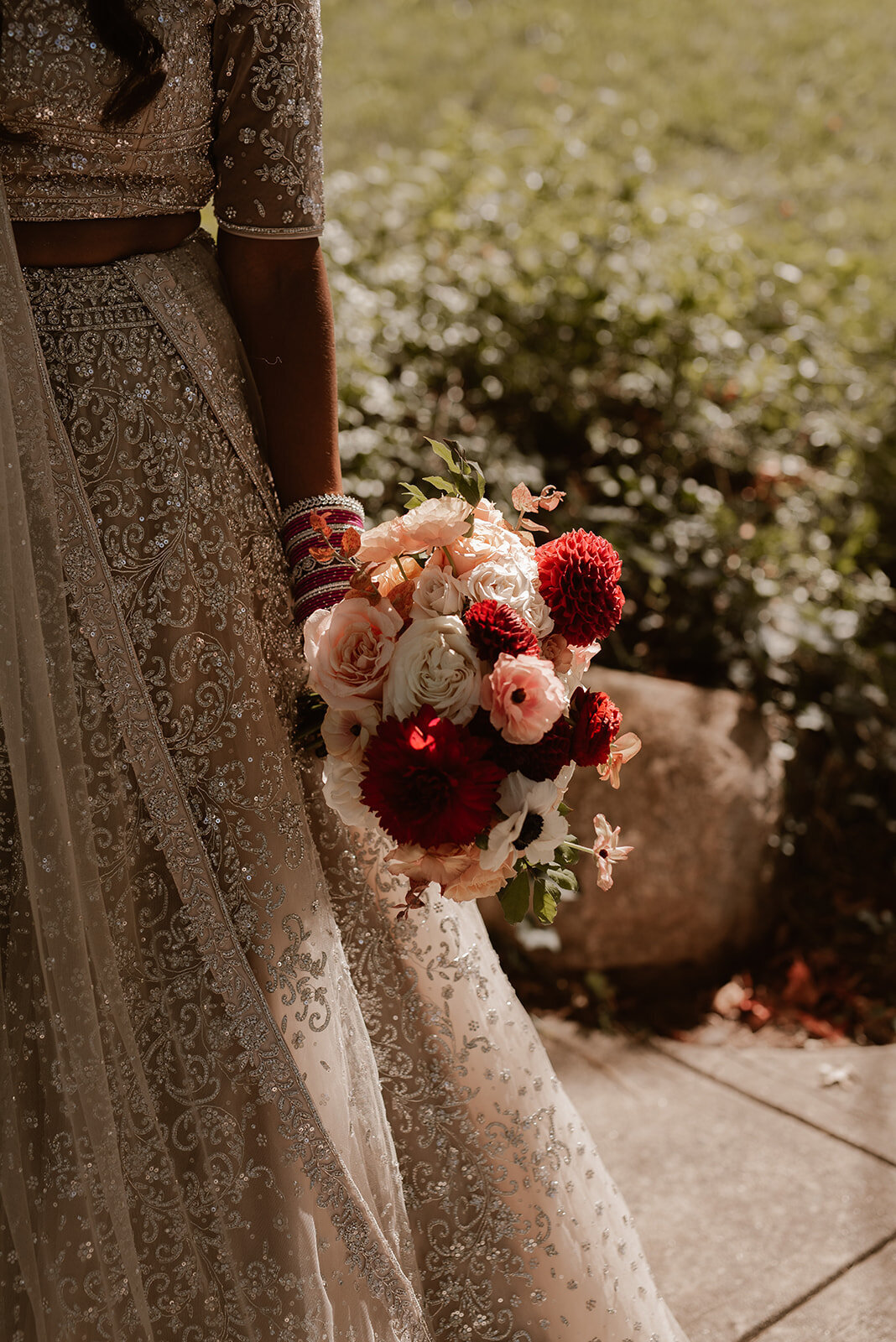 The bride gracefully walking outdoors, holding a bouquet of blush and deep red florals, her gown flowing behind her.