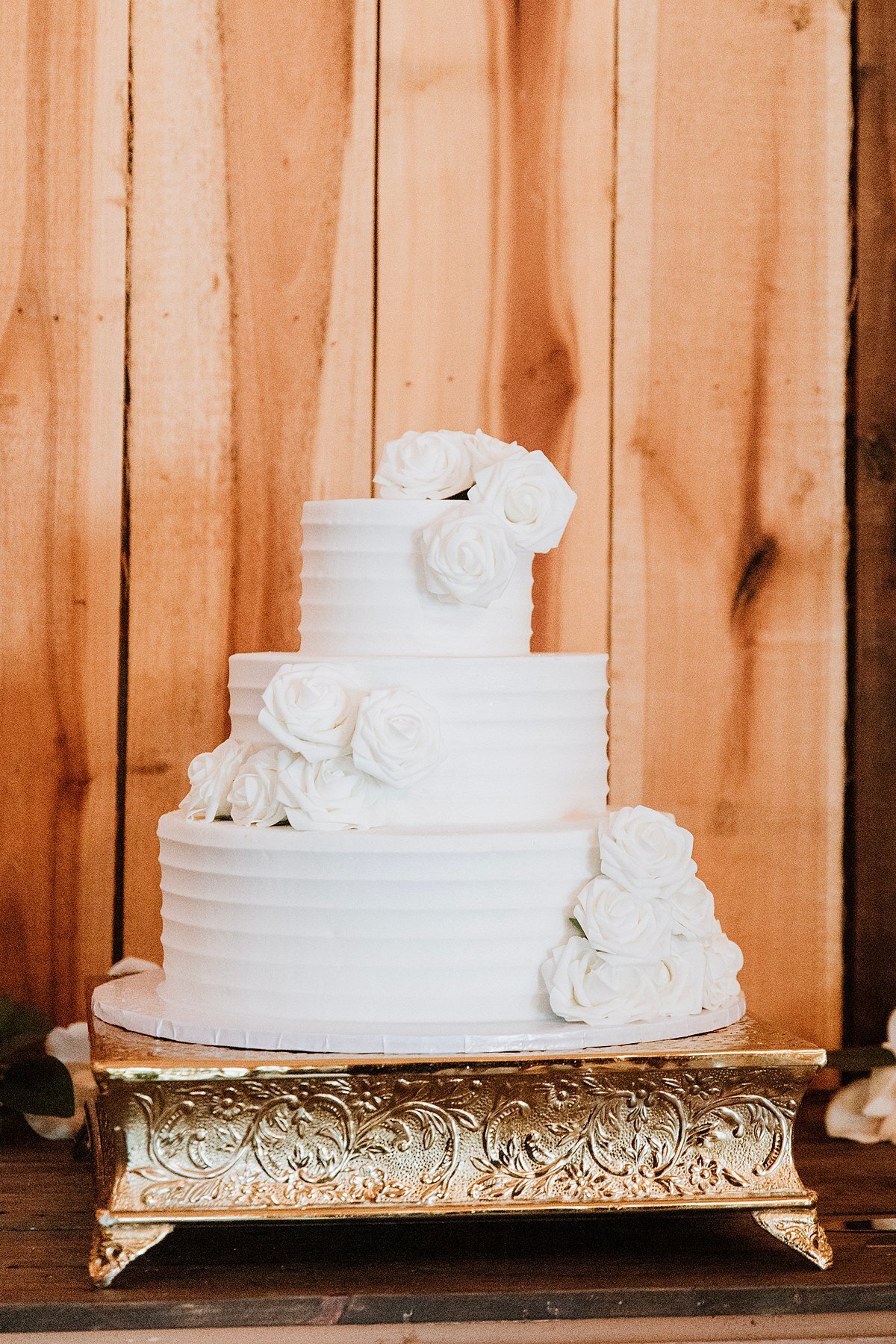 Elegant three tier white wedding cake with white flowers on a square silver cake stand against a light wood wall at Steel Magnolia Barn.