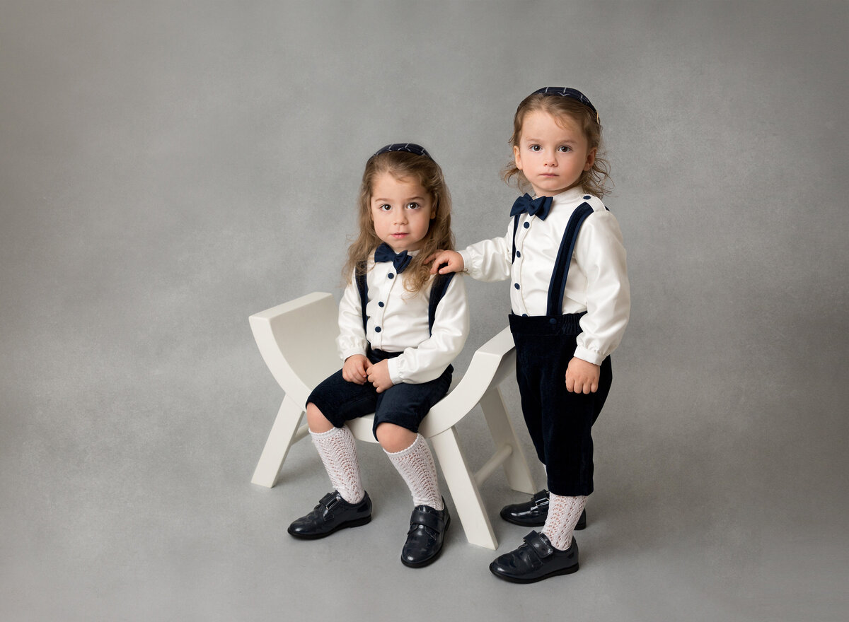 Toddler boys are posing for a milestone photoshoot. One boy is sitting in a chair, the other boy is standing with his hand on his brother's shoulder.