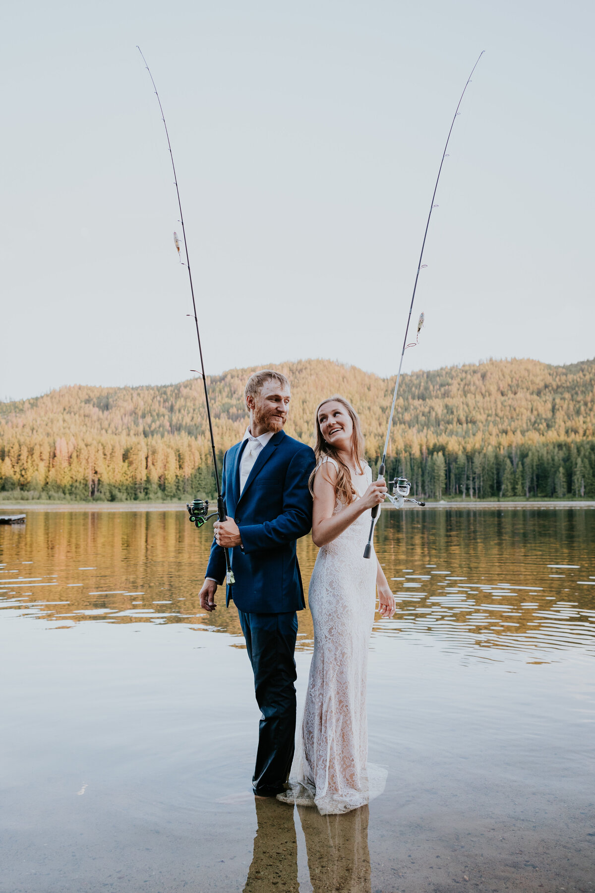 Bride and groom stand back to back on the shore of round lake, Idaho, while holding up their fishing poles.