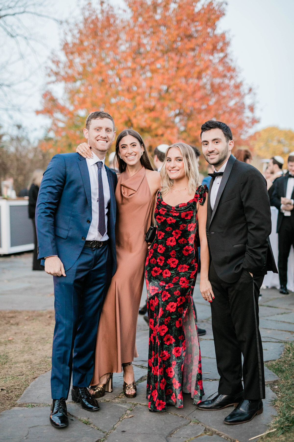 A group of four wedding guests, two men and two women, pose outdoors with a stunning autumn tree in the background