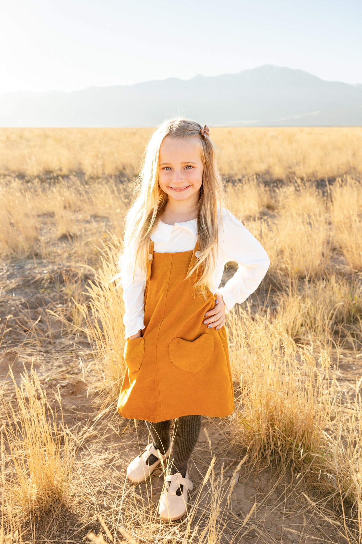 A young girl with long blonde hair stands confidently in a mustard-colored dress with heart-shaped pockets, with the mountains of Eagle Mountain, Utah, in the background.