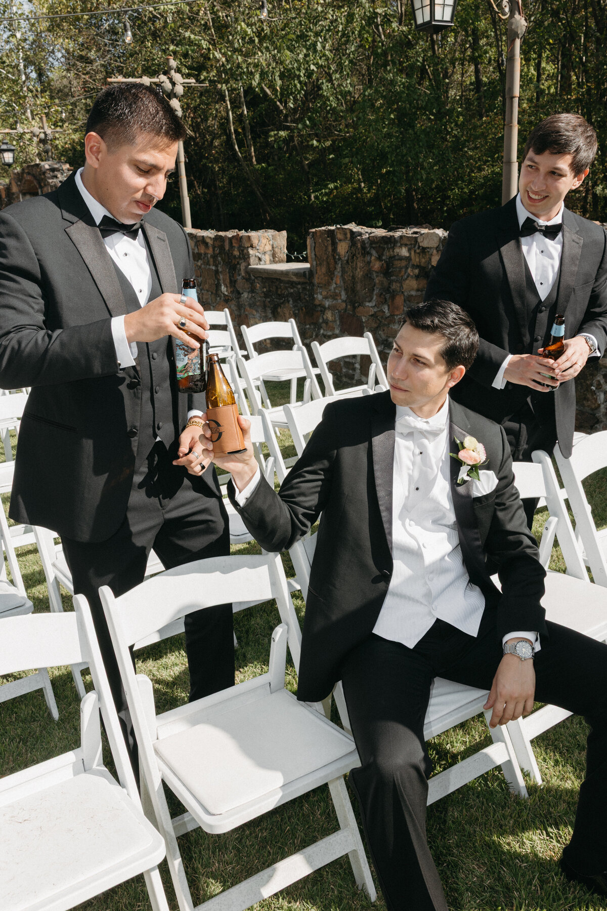 Groomsmen in classic black tuxedos sharing drinks before the outdoor ceremony, highlighting candid pre-wedding moments and refined boutonniere floral details.
