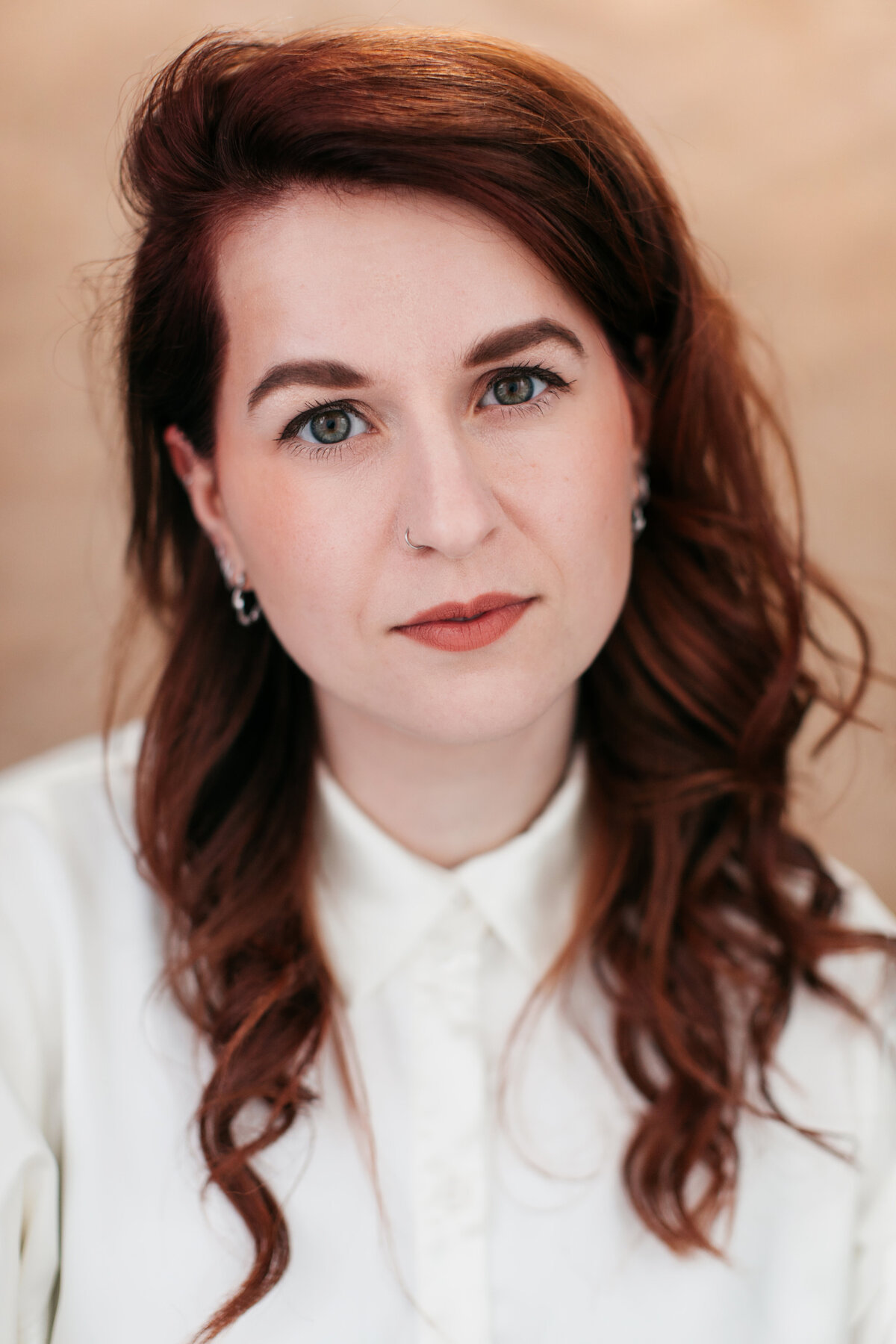 Castingfoto van vrouw met donkerrood haar in een witte blouse voor een beige achtergrond.