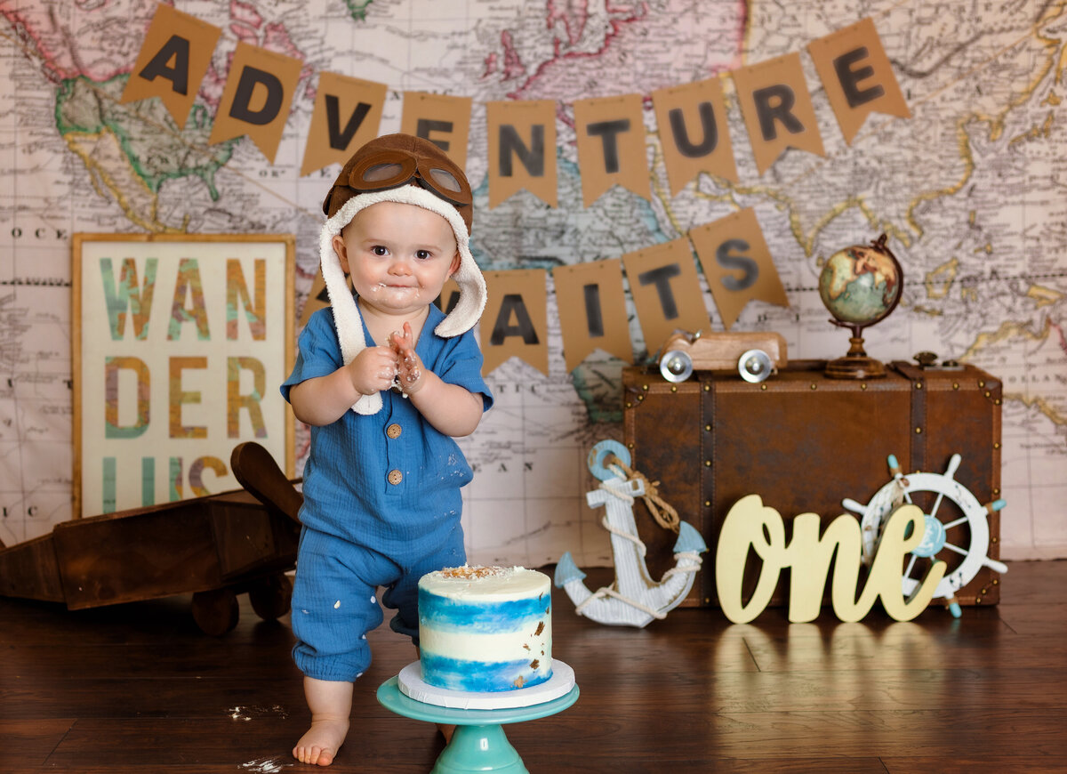 Cake Smash Photographer, a baby wears a pilot cap and stands before a map, and wanderlust backdrop for first birthday