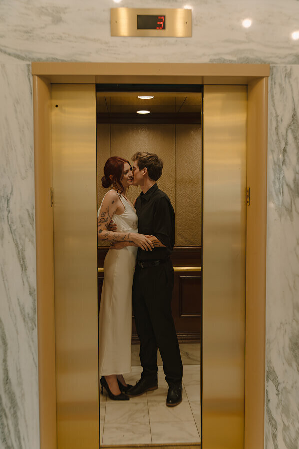 A man kisses his fiancé in an elevator. The doors are open as if you (the viewer) were peeking inside. 