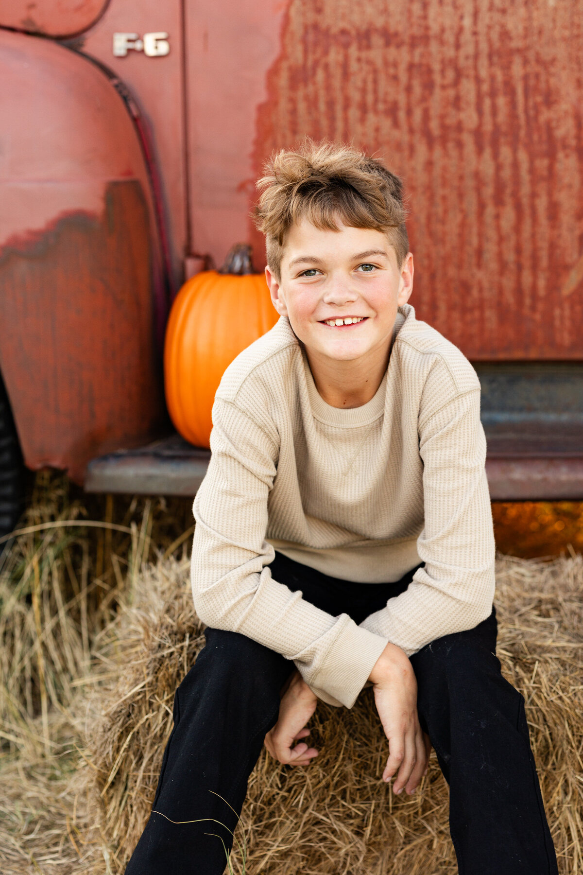 Preteen boy sits on a bale of hay in front of a vintage red farm truck and smiles at the camera.