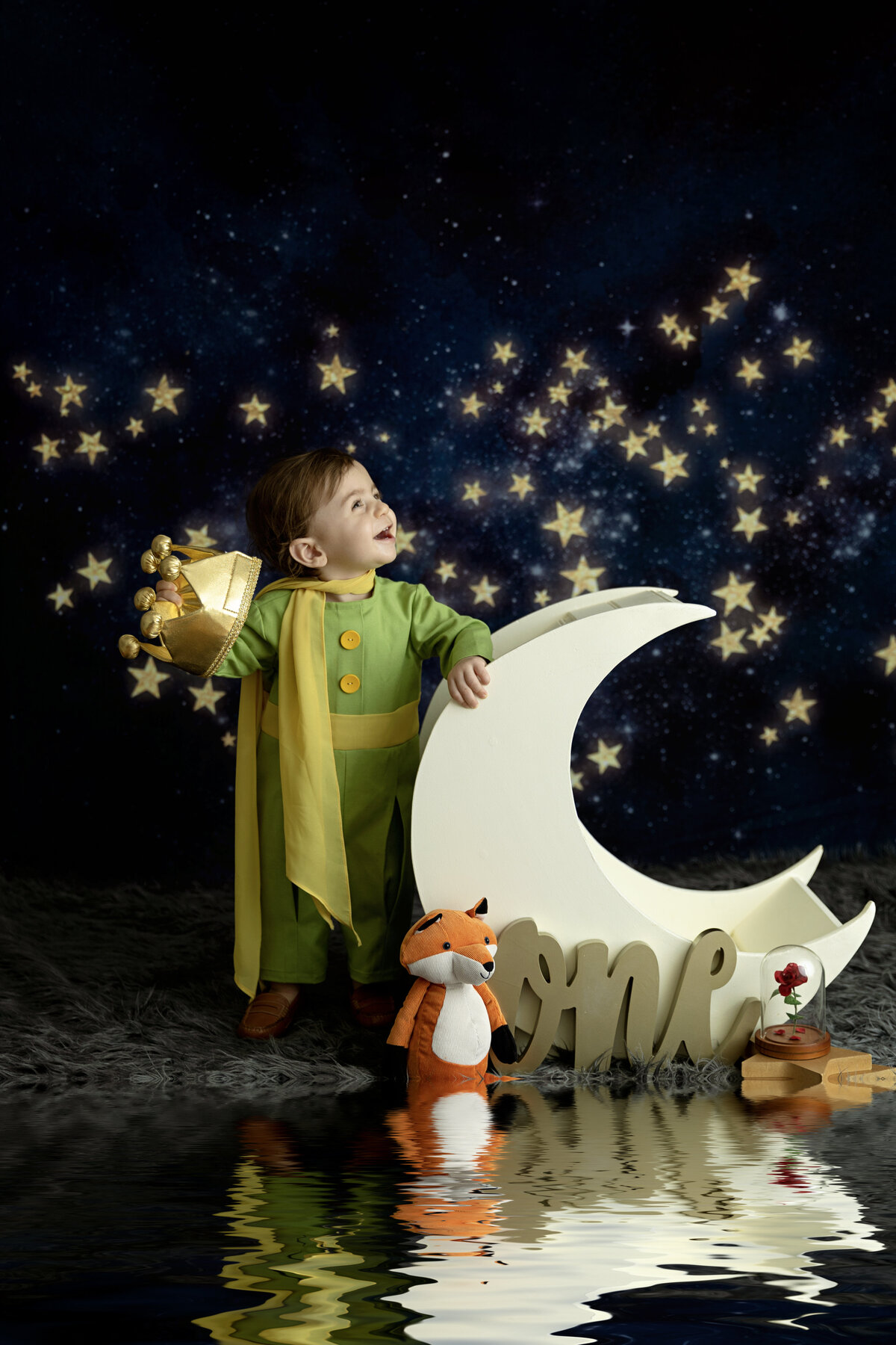 A toddler boy dressed as a prince for his first birthday leans against a moon shaped chair in a studio