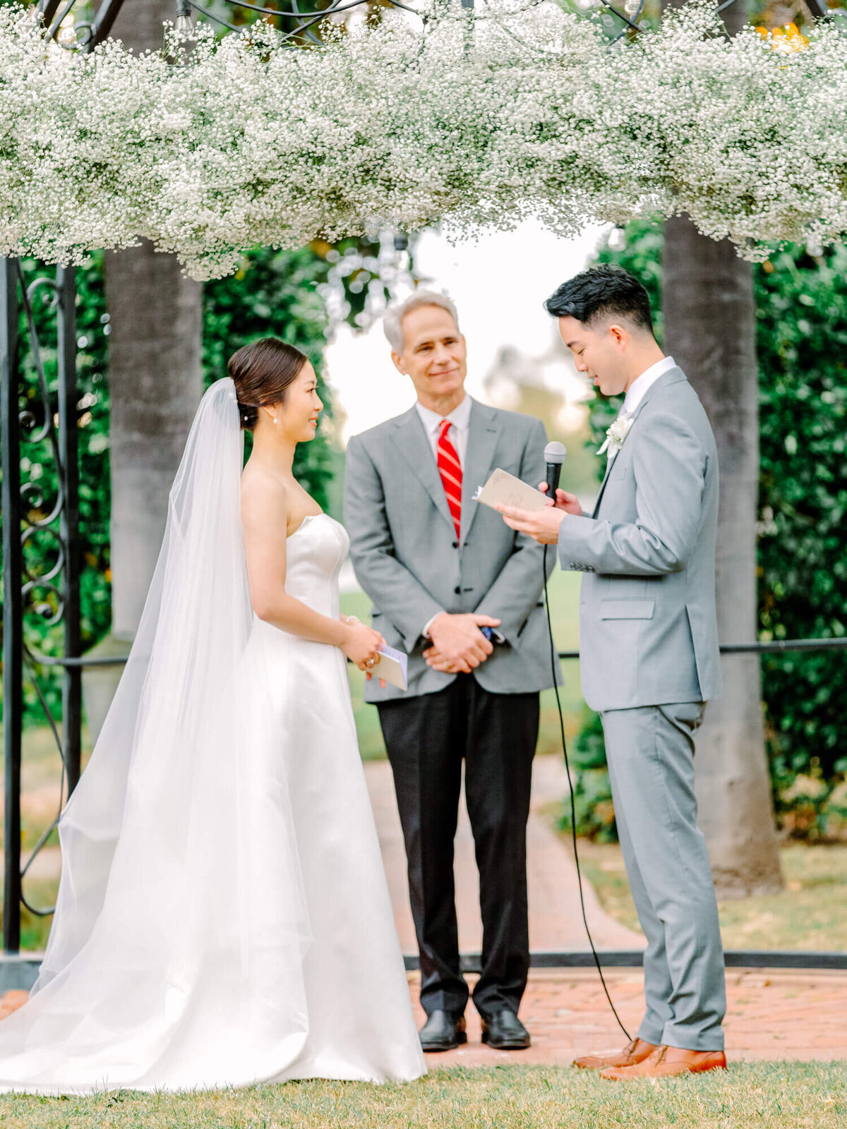 Bride and groom exchange vows under a white floral arch in an outdoor wedding ceremony. The officiant stands between them, creating a joyful atmosphere.