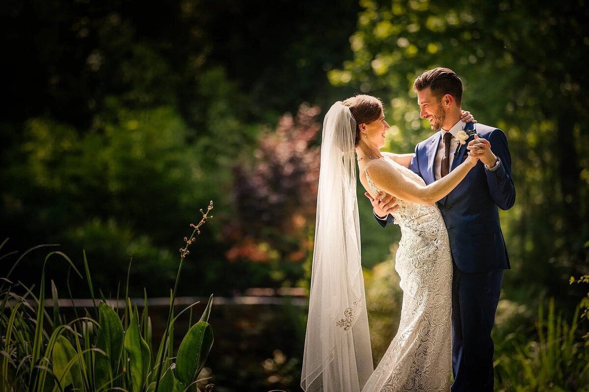 A bride and groom share a private dance in a serene garden, surrounded by lush greenery and bathed in sunlight.