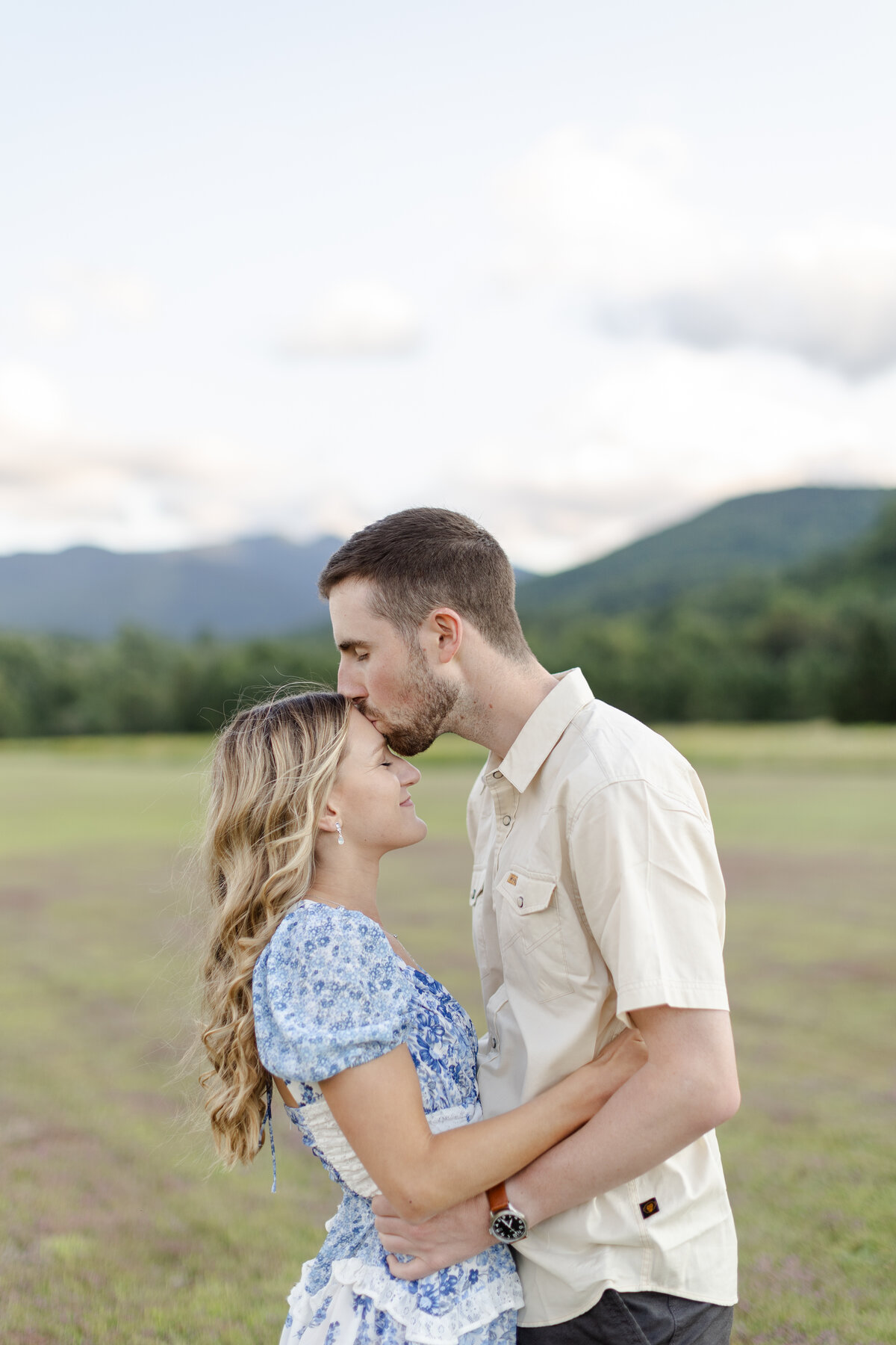 Engaged couple is standing in a field with their eyes closed while the groom kisses her forehead