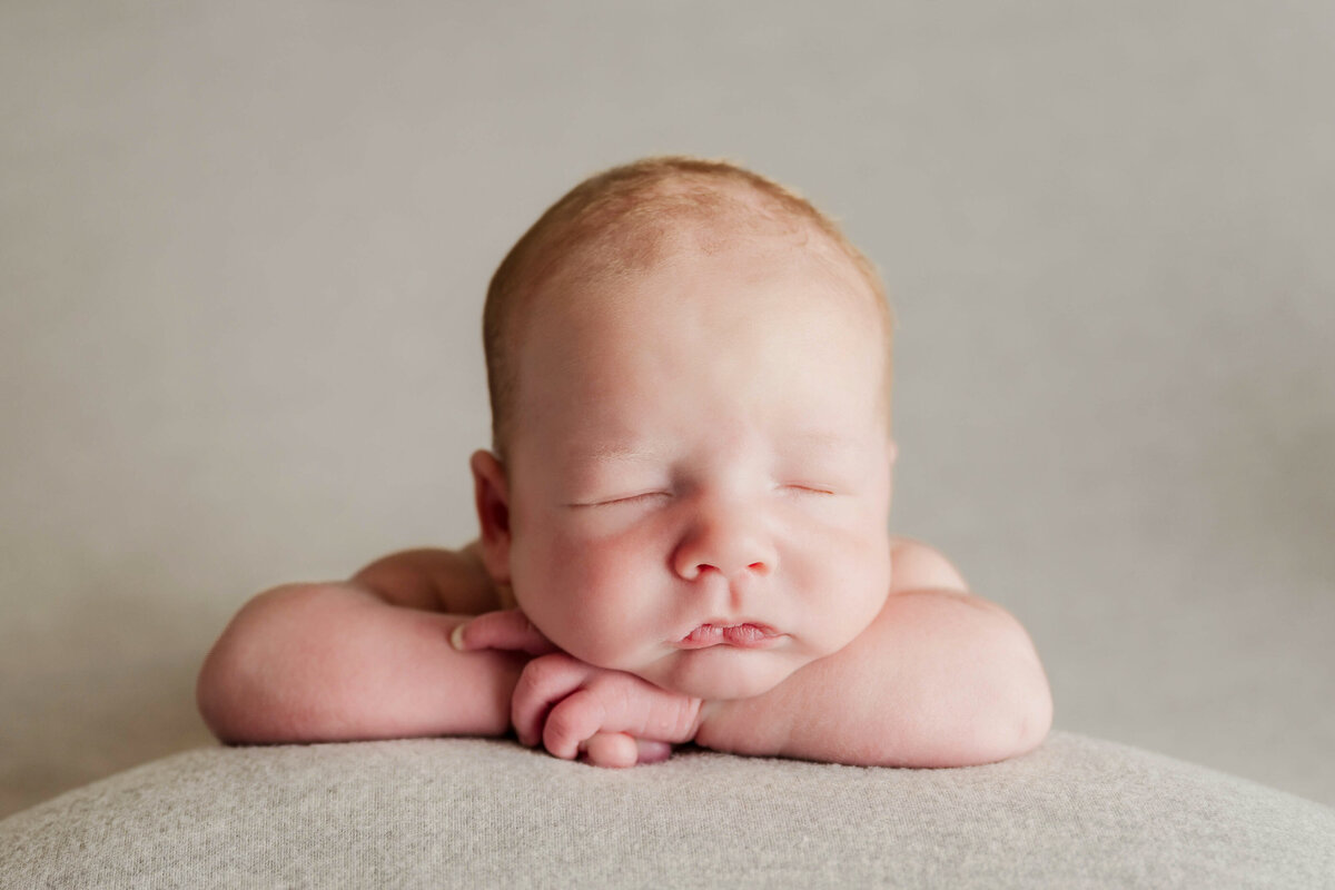 A newborn baby resting  his chin on his arms on a light beige backdrop in Raleigh NC photo studio.