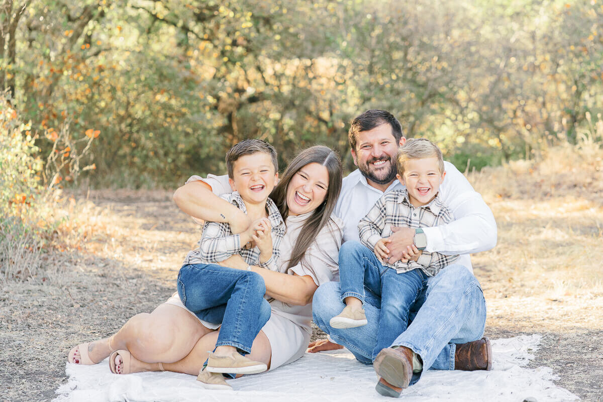 a family with two young boys sits on a white blanket and laughs for their photos with an Austin family photographer.