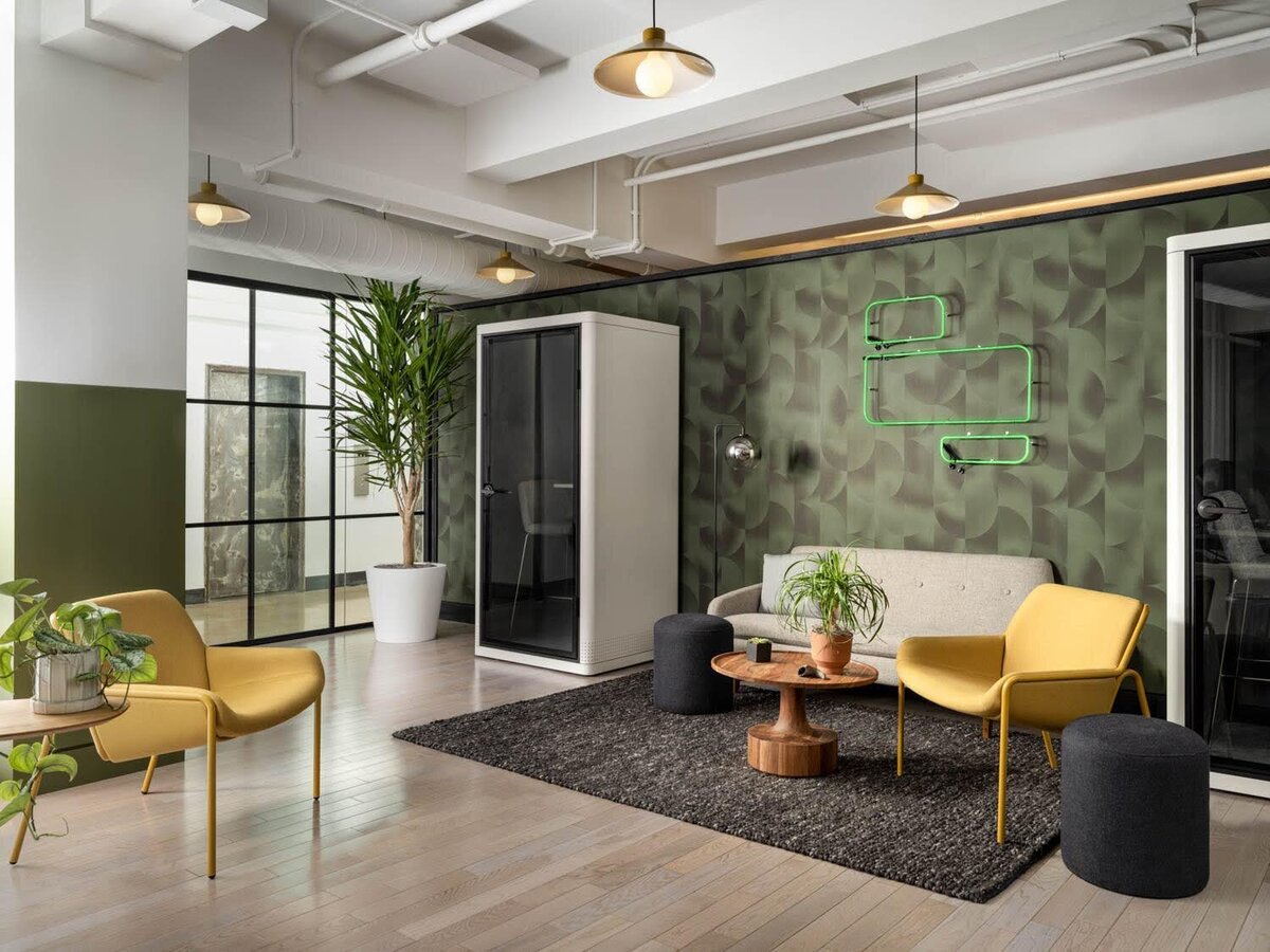 Open lounge area with olive green geometric wallpaper, mustard yellow lounge chairs, and a central acoustic phone booth.