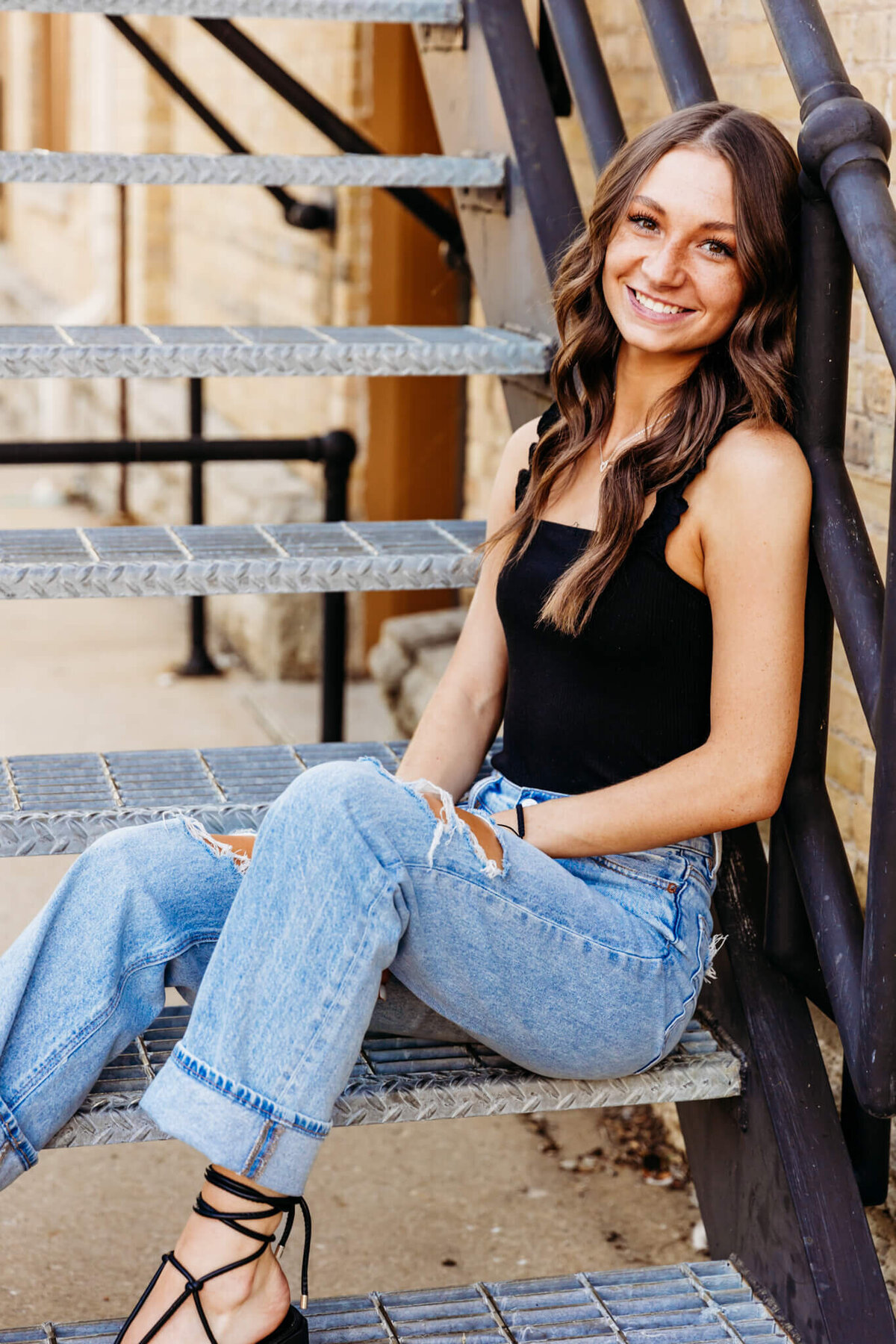 beautiful young woman in a black top and jeans sitting on a metal staircase and smiling captured by Ashley Kalbus Photography
