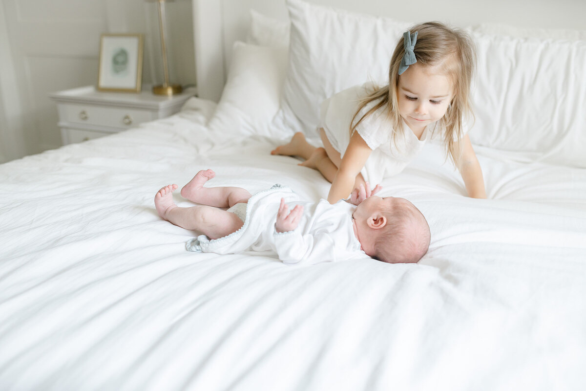 a little girl playing with her newborn sibling on a white bed