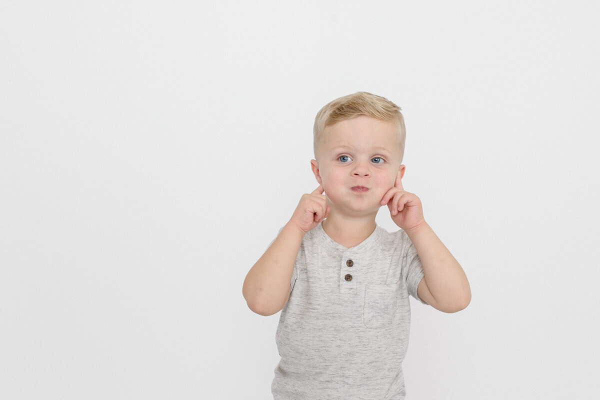 Close-up of a little boy with blonde hair pulling his cheeks with his hands and making a funny face, looking away from the camera in a white studio.