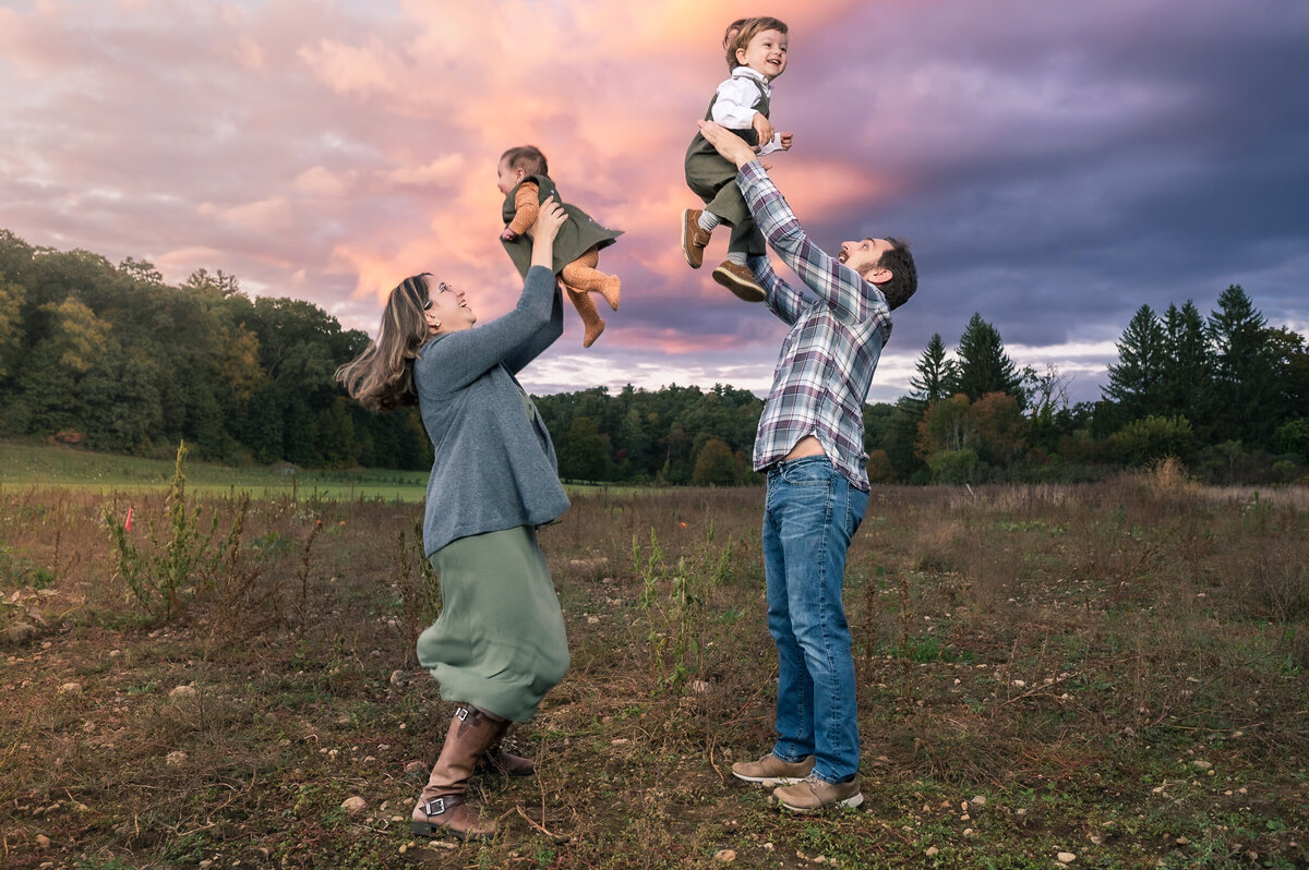 Mom and dad tossing kids in the air in a lincoln, ma pumpkin patch with a colorful sunset