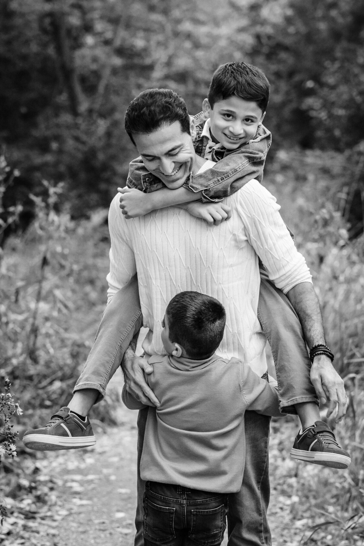 A black-and-white photo of a father carrying one son on his back while the other hugs him from the front. The father is smiling warmly, with a backdrop of trees and nature.