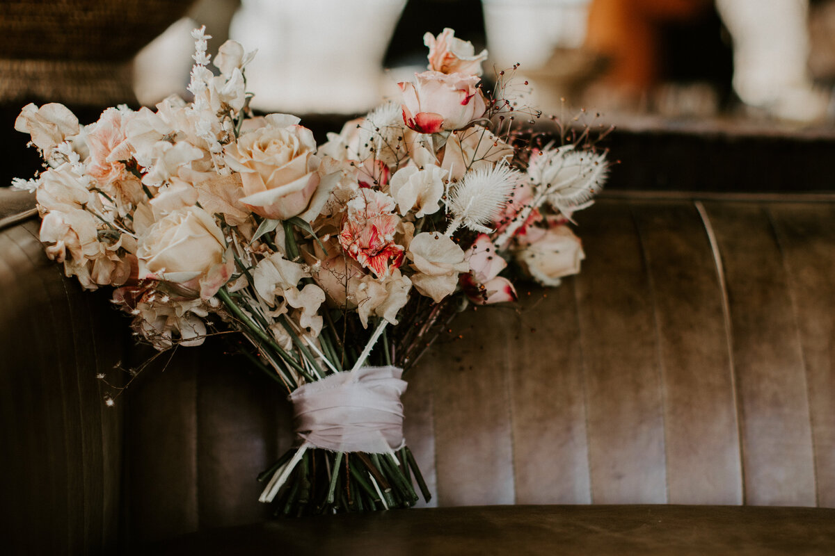 A bouquet of blush-colored flowers tied in ribbon placed against a brown leather coach.