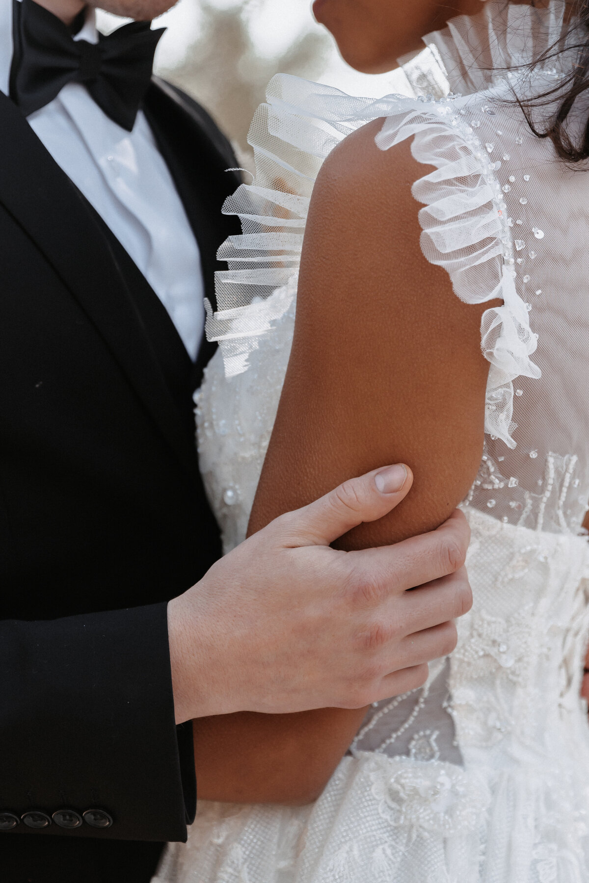 Close-up of groom's hand gently holding the bride's arm, highlighting her ruffled lace wedding gown.