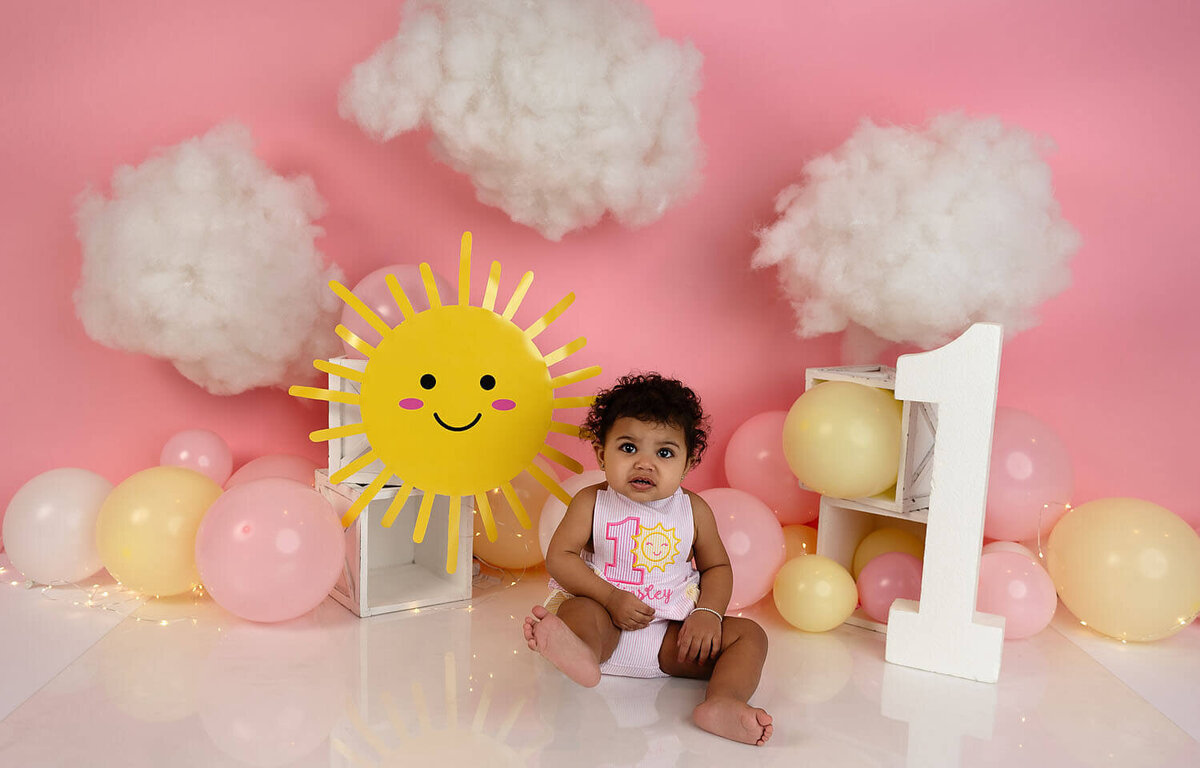 A young toddler girl sits in a happy studio set up in a first birthday shirt