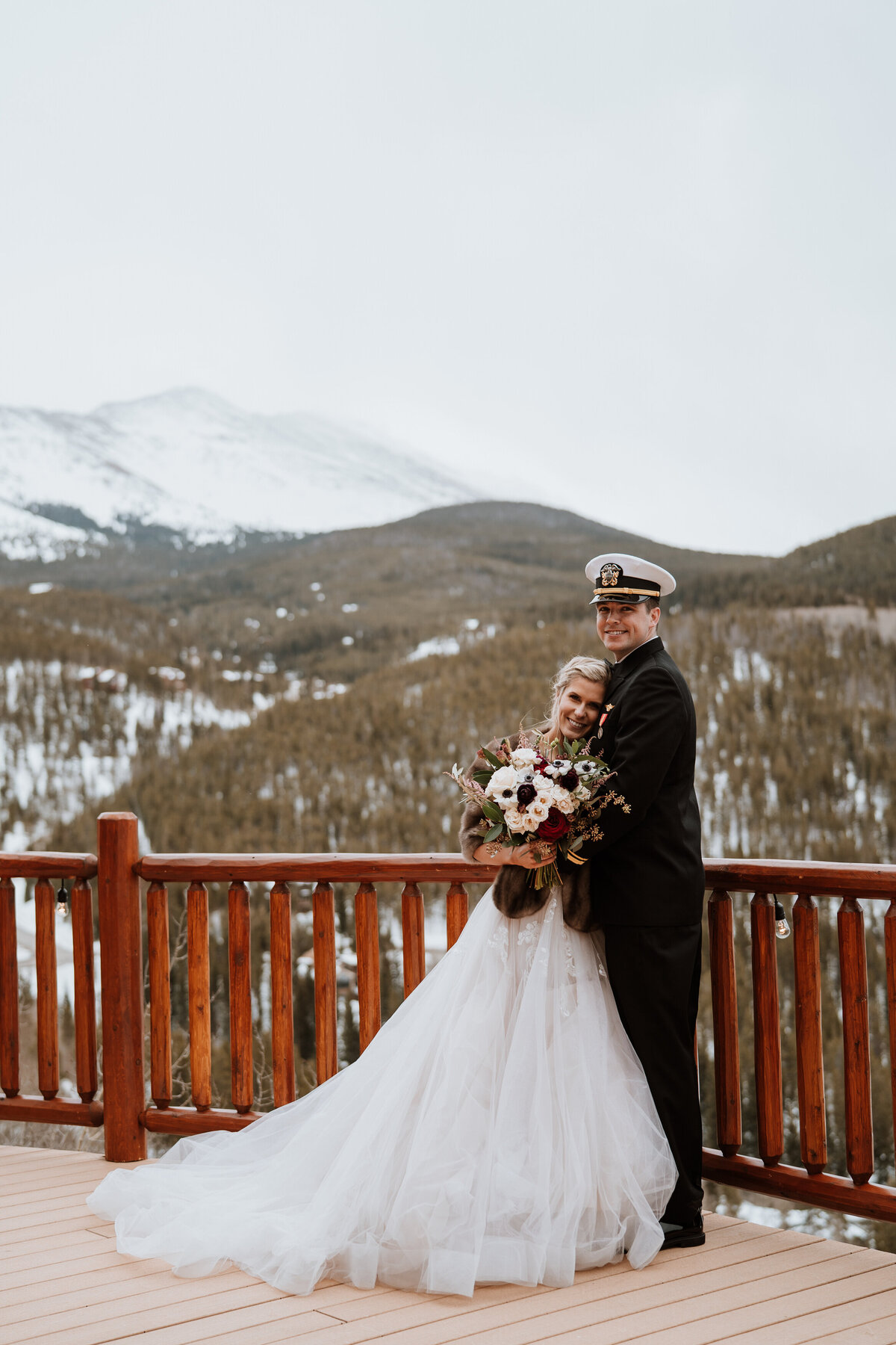 Navy groom and bride embrace on lodge deck with snowy Breckenridge mountain backdrop.